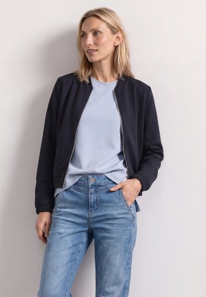 Femme aux cheveux blonds portant une veste noire zippée, un t-shirt bleu clair et un jean bleu, debout contre un mur blanc uni.