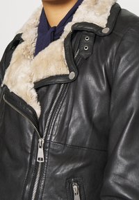 Giacca in pelle nera con colletto in pelliccia sintetica, chiusura con zip e tasche laterali. Presenta hardware argentato e superficie testurizzata.