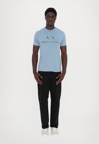 Gaiši zils īsām piedurknēm t-krekls ar melnu "ARMANI EXCHANGE" grafiku, kombinēts ar melnā krāsā taisna piegriezuma biksēm un baltajiem sporta apaviem.