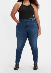 Tmavě modré vysoké džíny s přiléhavým střihem, s kapsami a zap ínáním na knoflík, v kombinaci s černým tank topem a botami na podpatku.