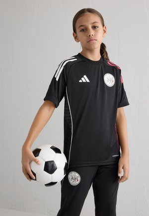 Zwarte voetbaluniform met witte Adidas-strepen, een ronde logo en rode accenten, gecombineerd met zwarte broek. Speler houdt een zwart-wit voetbal vast.