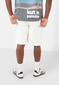 Shorts di denim bianco con orli sfilacciati, t-shirt grigia con grafica che presenta il testo "lost in paradise" e sneakers bianche a strisce nere.