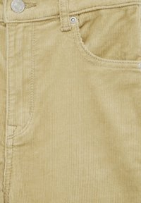 Détail d’un pantalon beige en velours côtelé montrant une poche avant, un passant de ceinture et un bouton métallique avec des surpiqûres.