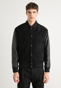 Black bomber jacket with suede body, leather sleeves, ribbed collar, and cuffs. Features snap button closure and embroidered logo on chest.