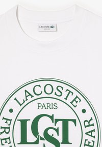 Camiseta de algodón blanca con un logo circular verde que incluye el texto "LACOSTE," "PARÍS" y "FRE EAR" en letras en negrita. Diseño de corte holgado.