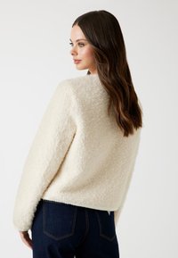 Donna con lunghi capelli castani che indossa un giubbino di pile testo crema e jeans blu scuro, di profilo su uno sfondo bianco semplice.