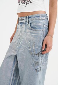 Lyseblå denimjeans med løst fit. Har sølvbroderi af en sommerfugl og et kranie på venstre ben, fem lommer og lukning med metalknap.