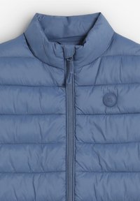 Chaqueta puff azul con cuello alto, diseño acolchado, cremallera frontal y un pequeño logo redondo en el pecho. Material suave y ligero.