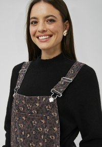 Salopette en denim motif léopard avec des clips métalliques réglables, associée à un pull en maille noir. Comprend une poche avant.