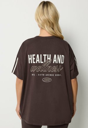 Blonde Person, die ein locker sitzendes dunkelbraunes T-Shirt mit weißem Text „HEALTH AND wellness NYC - FIFTH AVENUE 00981. 1998“ auf dem Rücken trägt.