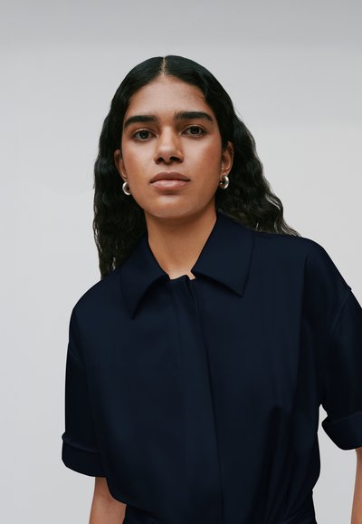 Camisa azul marino de cuello con una textura suave, de manga corta con los puños enrollados y cierre de botones en la parte delantera, usada por una persona con cabello rizado.