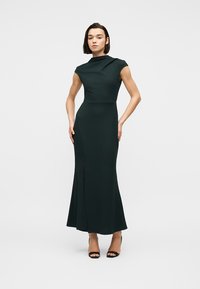 Γυναίκα που φοράει μαύρο αμάνικο φόρεμα μέχρι το πάτωμα με ψηλή λαιμόκοψη και μαύρα ψηλοτάκουνα παπούτσια ανοιχτά στα δάχτυλα, στέκεται μπροστά σε απλό φόντο.