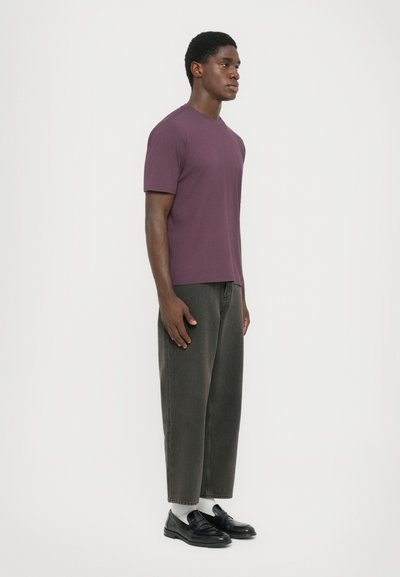 Man standing sideways wearing a purple short-sleeve shirt, cropped dark gray pants, white socks, and black loafers against a plain background.