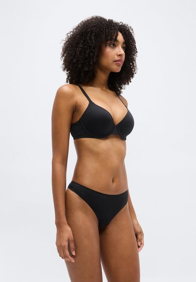 Νεαρή γυναίκα με σγουρά μαλλιά, φορώντας μαύρο σουτιέν και μαύρο εσώρουχο που ταιριάζει, στέκεται σε ουδέτερη στάση μπροστά σε απλό φόντο.
