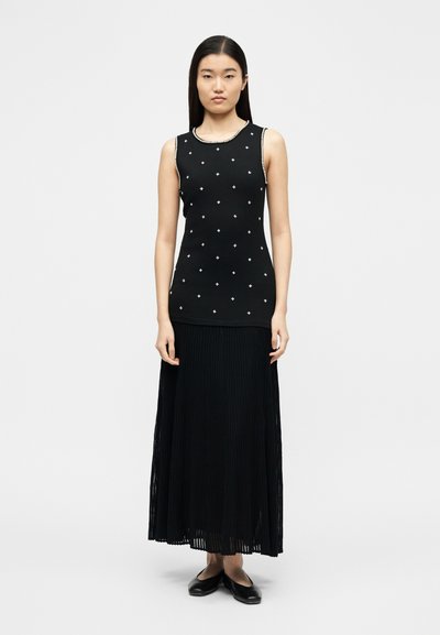 Γυναίκα που φοράει μαύρο αμάνικο τοπ με μικρά λευκά σχέδια και μακριά μαύρη πλισέ φούστα, συνδυασμένη με μαύρα φλατ παπούτσια.