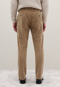 Braune Cordhosen mit gerippter Textur, zwei Gesäßtaschen mit Knöpfen und einem maßgeschneiderten Schnitt, kombiniert mit schwarzen Schuhen.