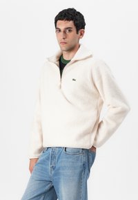 Crewneck pullover in cream fleece, featuring a half-zip collar and green logo detail. Paired with light blue denim jeans.