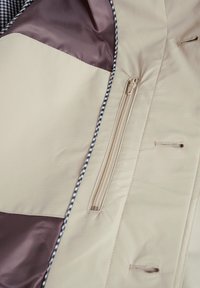 Bež in rjav plašč z glatko, gladko teksturo, ki ima zadrgo in kontrastno karirano podlogo ob ovratniku.