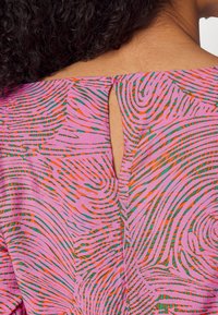 Primo piano della schiena di una persona con capelli ricci che indossa un top rosa con motivi astratti a spirale verdi e arancioni e una piccola apertura a forma di chiave vicino al collo.