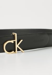 Čierny kožený opasok s zlatou kovovou prackou s logom "ck". Hladký povrch s embosovaným brandingom "Calvin Klein" na páske.