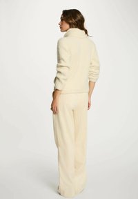 Maglione a coste di colore crema con colletto alto e maniche arrotolate, abbinato a pantaloni ampi di tessuto testurizzato in crema. Tessuto morbido e caldo.