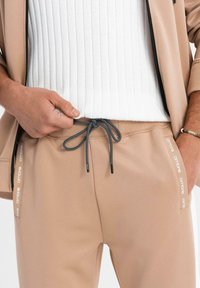 Personne portant un pantalon de survêtement beige avec un cordon noir et une veste zippée beige, tirant un pull blanc côtelé vers le haut.