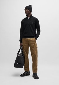 Zwarte rits-trui, tan cargo broeken, zwarte combat boots en een zwarte tas. Beanie muts. Focus op casual, functionele ontwerpelementen.