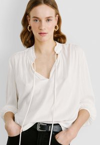Blusa bianca con colletto arricciato, scollo a V e cordini. Le maniche lunghe presentano polsini elasticizzati. Tessuto leggero e morbido. Cintura nera.