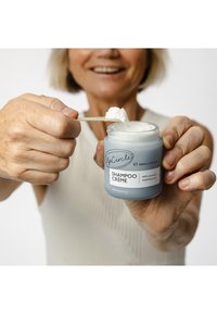 Jasnoniebieski słoik z napisem "Shampoo Crème", z białym tekstem i teksturowaną pokrywką, trzymany przez dłoń z aplikatorem białego kremu.