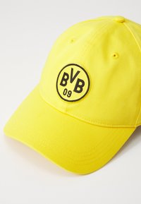 Żółta czapka bejsbolowa z zaokrągloną koroną, ozdobiona czarnym okrągłym logo z napisem "BVB 09" na środku. Wykonana z tkaniny o teksturowanej powierzchni.