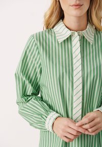 Zeleno-bílý pruhovaný košile s dlouhými rukávy, límcem na knoflíky a elastickými manžetami. Vertikální pruhy se střídají v šířce a směru.