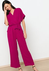 Fuchsia wijde jumpsuit met korte mouwen, top in wikkelstijl en zelfbindende riem. Soepele textuur, ontspannen pasvorm en minimalistisch ontwerp.
