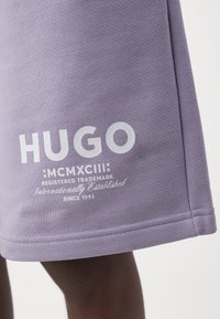 Gros plan d’un vêtement lavande avec un texte blanc indiquant « HUGO MCMXCIII REGISTERED TRADEMARK Internationally Established SINCE 1993 » près de l’ourlet.