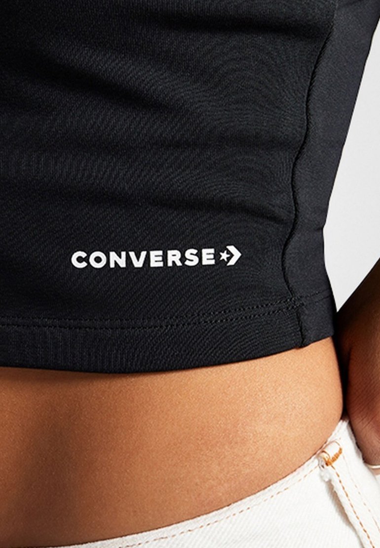 Camiseta corta negra hecha de tela elástica, con un logo blanco de "Converse" y un acento de estrella cerca del dobladillo. Textura suave.