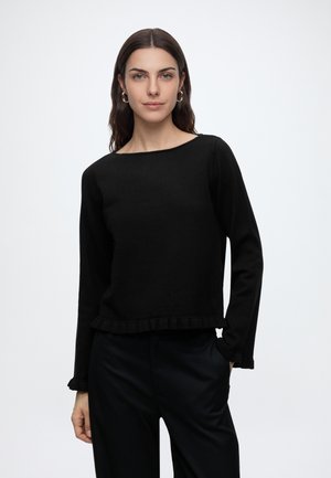 Femme aux longs cheveux foncés portant des boucles d’oreilles créoles dorées et un haut noir à manches longues avec des bords froncés, debout avec une main dans la poche.