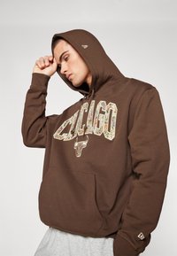 Hnedá mikina s kapucňou s nápisom "CHICAGO" a grafickým motívom býka v mnohobarevnom kvetinovom dizajne. Obsahuje predné vrecko a rebrované manžety.