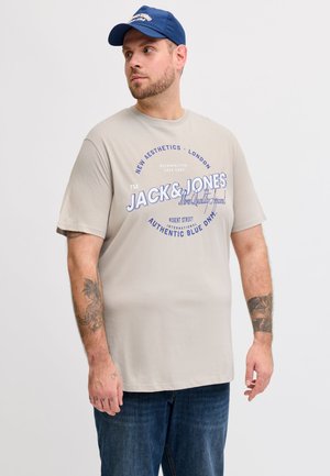 Bärtiger Mann trägt ein beiges Jack & Jones T-Shirt mit blauem Text und eine blaue Kappe, steht vor einem schlichten weißen Hintergrund.