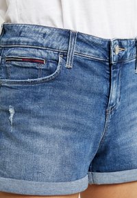 Pantalones cortos de mezclilla en azul medio con dobladillo deshilachado, ligeros desgastes y un dobladillo doblado. Presenta un diseño estándar de cinco bolsillos y herrajes de metal.