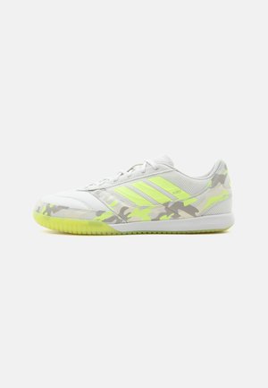Zapatilla deportiva blanca de corte bajo con detalles en camuflaje amarillo neón y tres franjas amarillas en el lateral, diseñada para uso en interiores.