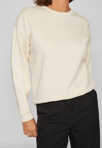 Crème-kleurige sweatshirt met lange mouwen, een ronde halslijn en geribde manchetten. De stof lijkt glad en heeft een relaxte pasvorm. Gekenmerkt door zwarte broeken.