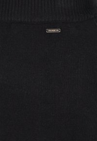 Schwarzer gestrickter Stoff mit geripptem Textur am oberen Rand und einem kleinen Metalletikett mit der Aufschrift "DRIMASTER", das an der Seite angenäht ist.