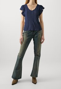 Maglietta corta blu navy con un motivo testurizzato, scollo a V e maniche a volant, abbinata a jeans svasati sbiaditi e tacchi a punta.