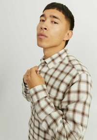 Camisa de franela de cuadros marrones y blancos con cuello abotonado, mangas largas y tela texturizada. El modelo ajusta el cuello mientras está de perfil.