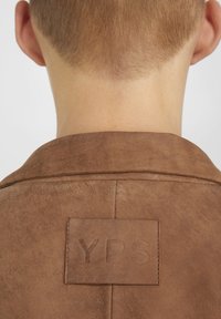 Zadní část krku a hlavy osoby s krátkými vlasy, která má na sobě hnědou bundu s našitou obdélníkovou záplatou s vyraženým nápisem "YPS".