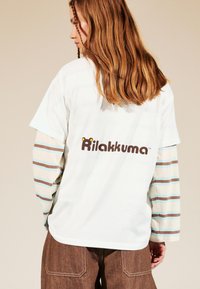 Mladý človek s dlhými hnedými vlasmi, oblečený v bielej a pruhovanej košeli s dlhým rukávom a hnedých nohaviciach, na ktorých chrbte je nápis 'Rilakkuma'.