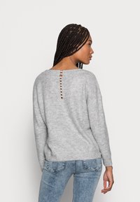 Grau gestrickter Pullover mit langen Ärmeln und scoop Rückseite, verziert mit einem dekorativen Knopfdetail entlang der Wirbelsäule. Kombiniert mit blauen Jeans.