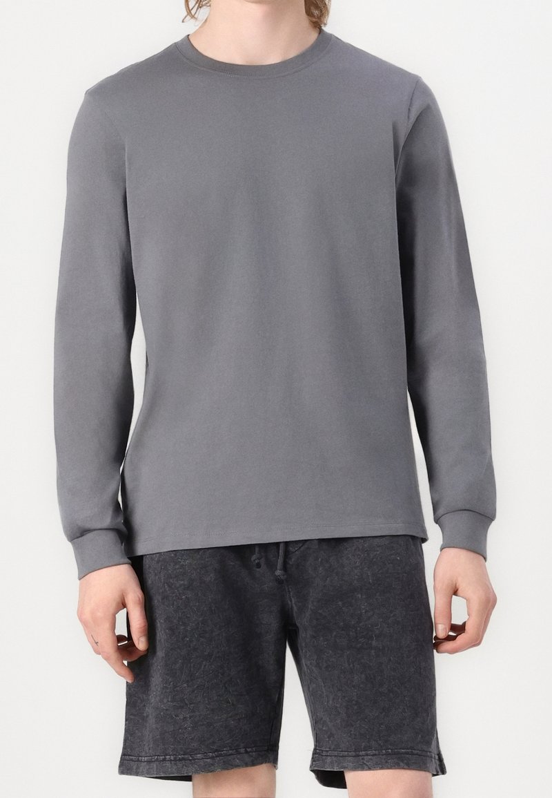 Mann trägt ein schlichtes graues Langarmshirt und schwarze gewaschene Baumwoll-Shorts mit Kordelzug, steht vor einem weißen Hintergrund.