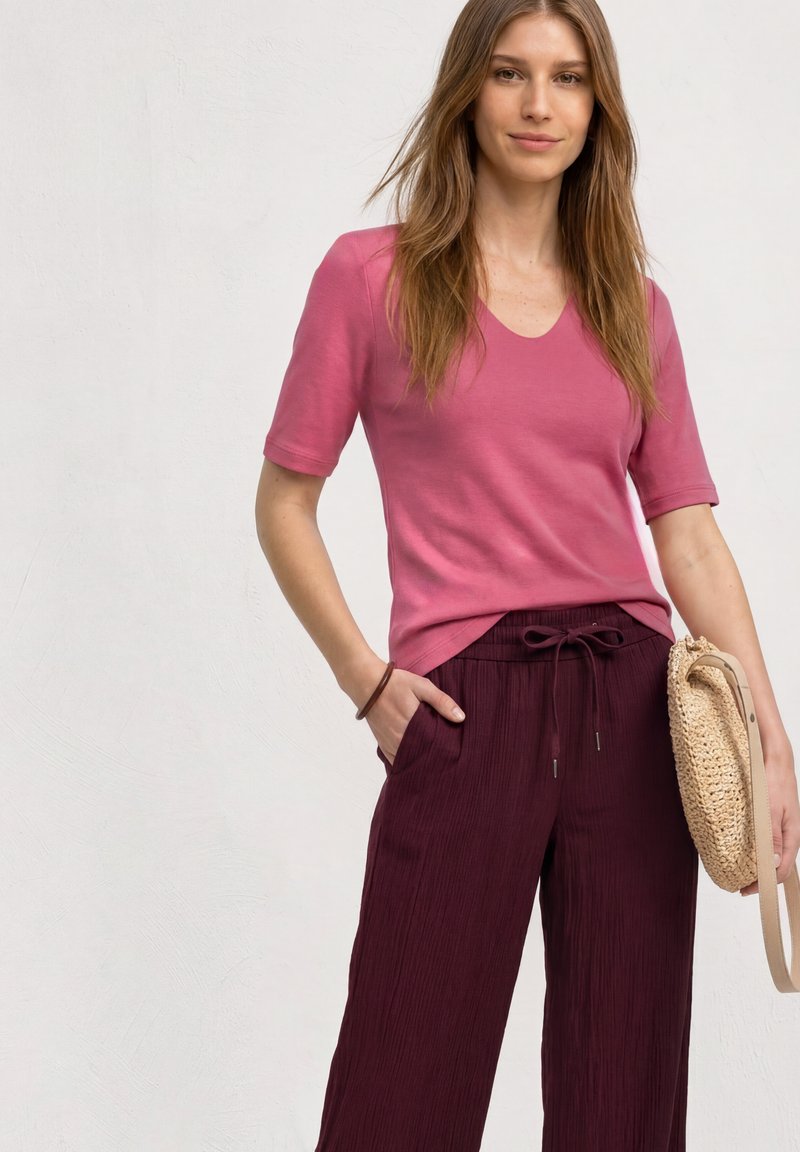 Femme portant un haut rose à manches courtes et un pantalon violet foncé, tenant un sac à main beige tissé, une main dans la poche, sur fond uni.