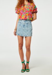 Barevná halenka s volánkovými rukávy, zdobená květinovým vzorem v růžové, zelené a modré barvě, v kombinaci s světle modrou denimovou sukní a zelenými podpatky.