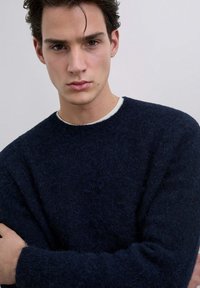 Navy blue wool sweater with a crew neck and long sleeves. Soft, textured fabric with a relaxed fit. White inner collar detail visible.